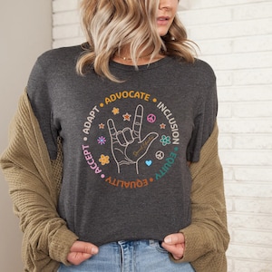 May include: A dark gray t-shirt with a circle design featuring the words "Accept, Adapt, Advocate, Equality, Equity, Inclusion" in a circular pattern around a hand making the American Sign Language symbol for "I love you".