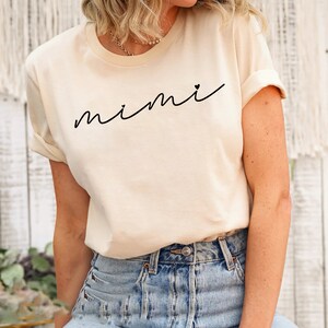 May include: A cream colored t-shirt with the word "mimi" written in black cursive with a small heart next to it.