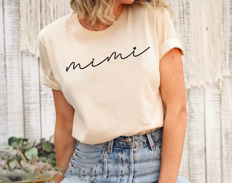 May include: A cream colored t-shirt with the word "mimi" written in black cursive with a small heart next to it.