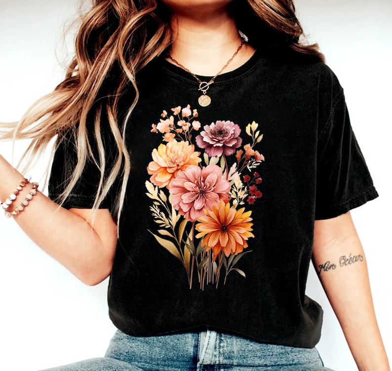 May include: A black short-sleeved t-shirt featuring a central graphic print of a colorful floral bouquet. The design includes various flowers in shades of orange, pink, and muted red, accompanied by green foliage and stems, centered on the chest.