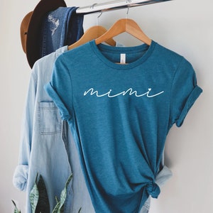 May include: A teal blue t-shirt with the word "mimi" written in white cursive with a small white heart after the "i".