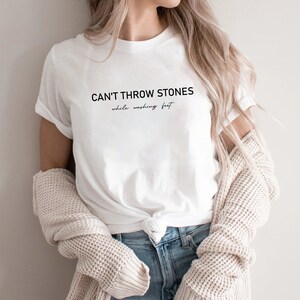 Puede incluir: Camiseta blanca con el texto "CAN'T THROW STONES while washing feet" impreso en la parte delantera.
