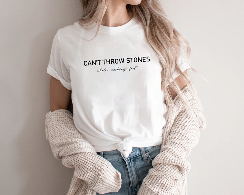 Puede incluir: Camiseta blanca con el texto "CAN'T THROW STONES while washing feet" impreso en la parte delantera.