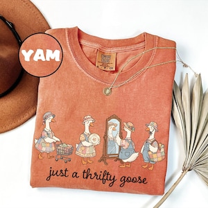 May include: A coral-colored Comfort Colors t-shirt featuring a whimsical illustration of geese dressed in vintage clothing. The phrase "just a thrifty goose" is printed below the geese. A gold necklace and a brown hat are also visible.