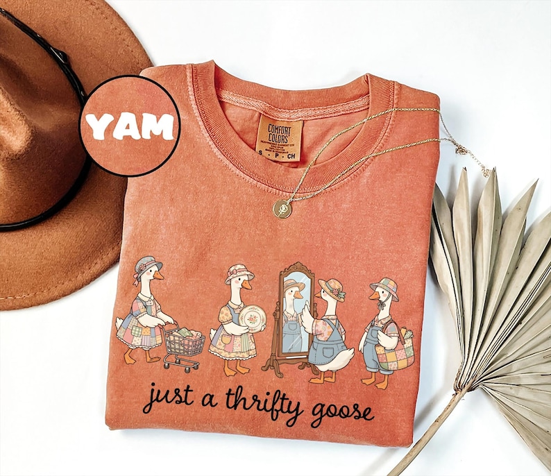 May include: A coral-colored Comfort Colors t-shirt featuring a whimsical illustration of geese dressed in vintage clothing. The phrase "just a thrifty goose" is printed below the geese. A gold necklace and a brown hat are also visible.