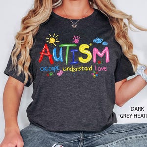 May include: Dark grey heather t-shirt with the word "AUTISM" in colorful embroidered letters. The shirt also features the words "accept understand love" and colorful graphics of a sun, clouds, and flowers.