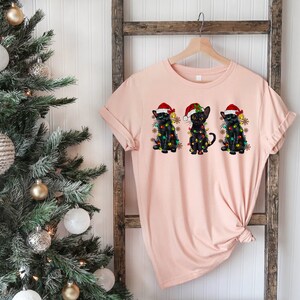 Pode incluir: Uma camiseta rosa com um desenho de tr&ecirc;s gatos pretos usando chap&eacute;us de Papai Noel e luzes de Natal. Os gatos est&atilde;o sentados em fila.