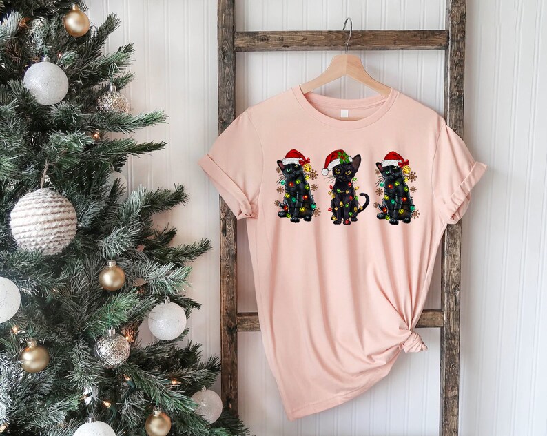 Pode incluir: Uma camiseta rosa com um desenho de tr&ecirc;s gatos pretos usando chap&eacute;us de Papai Noel e luzes de Natal. Os gatos est&atilde;o sentados em fila.