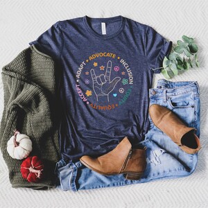 May include: A navy blue t-shirt with a graphic of a hand making the American Sign Language sign for "I love you". The graphic is surrounded by a circle with the words "Adapt", "Advocate", "Inclusion", "Equality", "Accept", "Unity" in a colorful font.