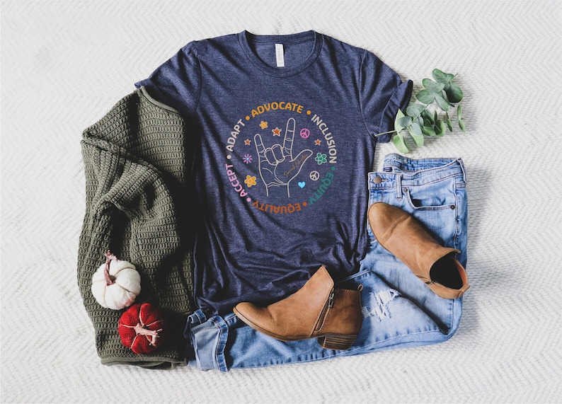 May include: A navy blue t-shirt with a graphic of a hand making the American Sign Language sign for "I love you". The graphic is surrounded by a circle with the words "Adapt", "Advocate", "Inclusion", "Equality", "Accept", "Unity" in a colorful font.