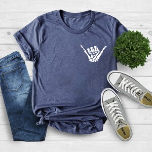 Op de afbeelding: Een marineblauw t-shirt met een witte skelethand die een shaka-teken maakt. Het shirt ligt op een wit houten oppervlak met een paar grijze Converse-schoenen en een blauwe jeans.