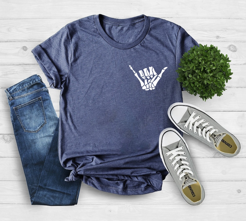 Op de afbeelding: Een marineblauw t-shirt met een witte skelethand die een shaka-teken maakt. Het shirt ligt op een wit houten oppervlak met een paar grijze Converse-schoenen en een blauwe jeans.