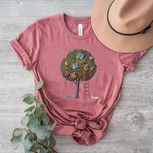 Pu&ograve; includere: Una maglietta rosa con una grafica di una persona che sale su una scala per raggiungere dei libri in un albero. L'albero &egrave; verde e i libri sono colorati.