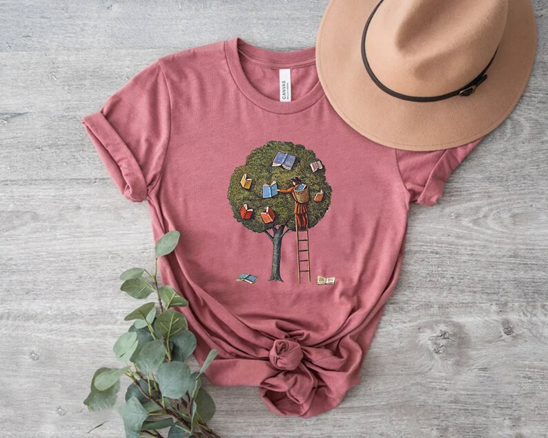 Pu&ograve; includere: Una maglietta rosa con una grafica di una persona che sale su una scala per raggiungere dei libri in un albero. L'albero &egrave; verde e i libri sono colorati.