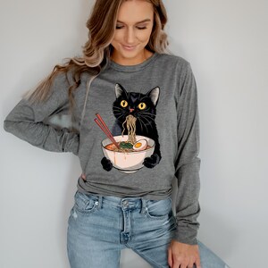 May include: A gray long-sleeved shirt with a black cat eating ramen noodles from a bowl. The cat has yellow eyes and is wearing a red headband.