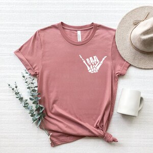 Op de afbeelding: Een stoffig roze T-shirt met een witte skelethand die een shaka-teken maakt.