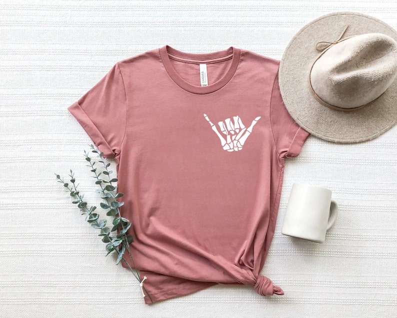 Op de afbeelding: Een stoffig roze T-shirt met een witte skelethand die een shaka-teken maakt.