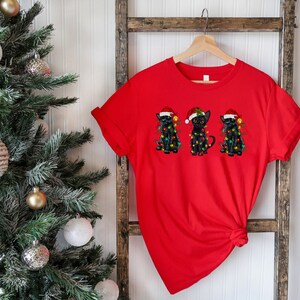 Pode incluir: Camiseta vermelha com um gr&aacute;fico de tr&ecirc;s gatos pretos usando chap&eacute;us de Papai Noel e luzes de Natal.
