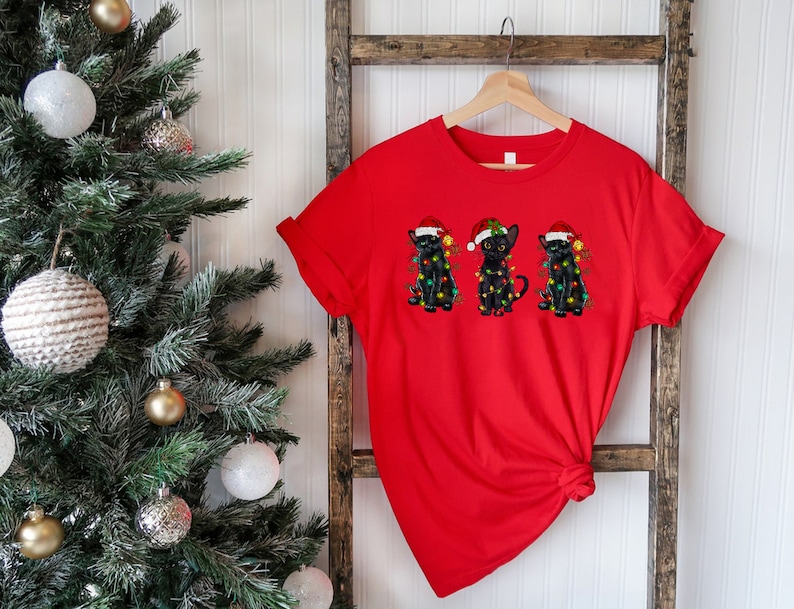 Pode incluir: Camiseta vermelha com um gr&aacute;fico de tr&ecirc;s gatos pretos usando chap&eacute;us de Papai Noel e luzes de Natal.