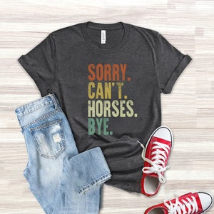 May include: A dark gray t-shirt with the text "SORRY. CAN'T. HORSES. BYE." in a vintage style font with colorful accents. The shirt is laid flat on a light wood surface with a pair of blue jeans and red sneakers.