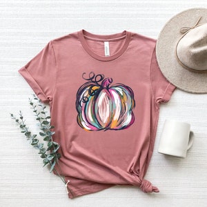 Może przedstawiać: T-shirt z kr&oacute;tkim rękawem w kolorze przybrudzonego r&oacute;żu z centralną grafiką przedstawiającą stylizowaną dynię. Ilustracja dyni wykonana jest za pomocą wyrazistych pociągnięć pędzla w odcieniach bieli, pomarańczu, turkusu i ciemnego błękitu.