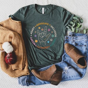 May include: A dark green t-shirt with a circle graphic featuring the words "Adapt, Accept, Advocate, Ally, Equity, Inclusion" and a hand making the American Sign Language sign for "I love you".