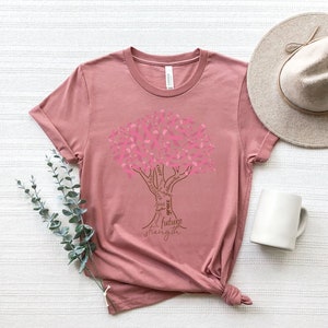 May include: A dusty rose t-shirt featuring a graphic of a tree where the leaves are composed of pink ribbons. The trunk and branches contain the words cure, hope, faith, grace, future, strength, fighter, and survivor in a brown script font.