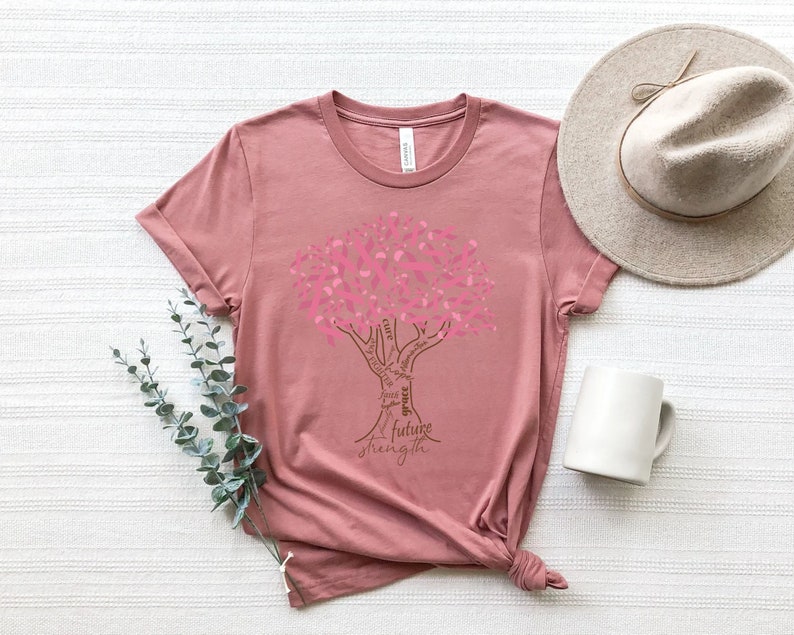 May include: A dusty rose t-shirt featuring a graphic of a tree where the leaves are composed of pink ribbons. The trunk and branches contain the words cure, hope, faith, grace, future, strength, fighter, and survivor in a brown script font.
