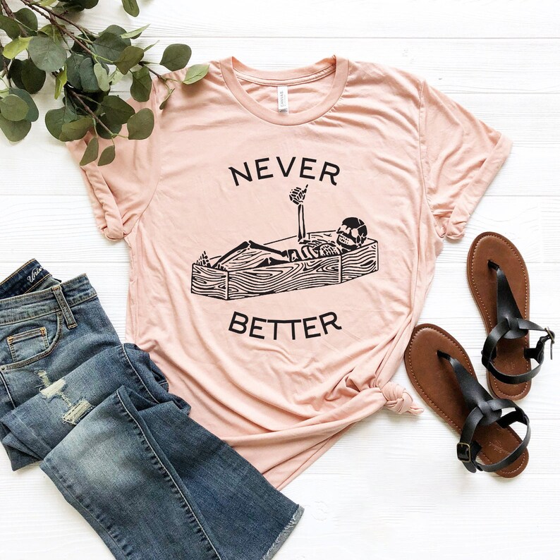 May include: A light pink t-shirt with a black skeleton graphic lying in a coffin with the text "NEVER BETTER".