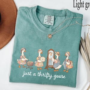 May include: Light green t-shirt with a crew neck and short sleeves. The shirt features a whimsical illustration of geese dressed in vintage clothing, with the text "just a thrifty goose" in a cursive font. A silver necklace with a pendant is draped across the shirt.