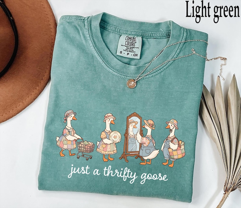 May include: Light green t-shirt with a crew neck and short sleeves. The shirt features a whimsical illustration of geese dressed in vintage clothing, with the text "just a thrifty goose" in a cursive font. A silver necklace with a pendant is draped across the shirt.