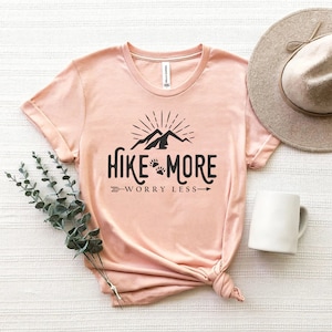 May include: A light pink t-shirt with the text "Hike More Worry Less" in black. The design includes mountains, paw prints, and an arrow. A beige hat and white mug are next to the shirt.
