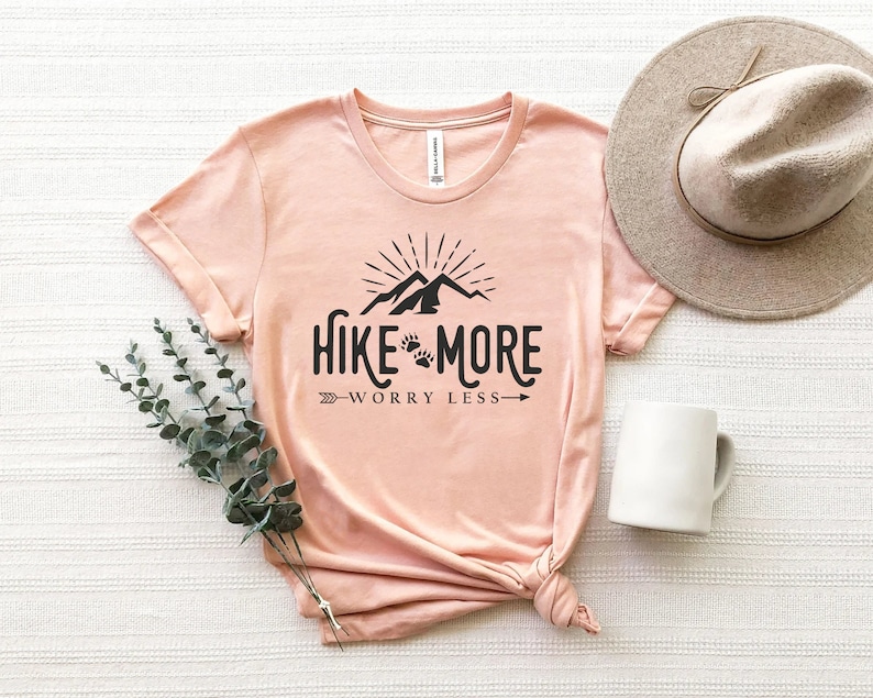 May include: A light pink t-shirt with the text "Hike More Worry Less" in black. The design includes mountains, paw prints, and an arrow. A beige hat and white mug are next to the shirt.