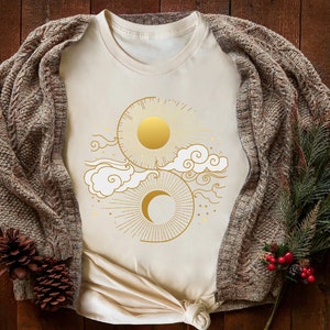 May include: A cream-colored t-shirt with a gold sun and moon design. The design features a sun with rays and a crescent moon with clouds and stars.