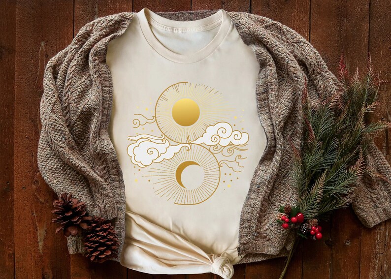 May include: A cream-colored t-shirt with a gold sun and moon design. The design features a sun with rays and a crescent moon with clouds and stars.