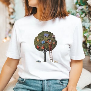 Pu&ograve; includere: Maglietta bianca con una grafica di una persona che sale su una scala per raggiungere un albero con libri come foglie. L'albero &egrave; verde e i libri sono colorati.