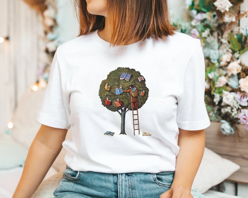 Pu&ograve; includere: Maglietta bianca con una grafica di una persona che sale su una scala per raggiungere un albero con libri come foglie. L'albero &egrave; verde e i libri sono colorati.