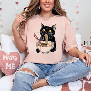 May include: A pink t-shirt with a black cat eating ramen noodles. The cat is holding a bowl of ramen with chopsticks. The text "Hug Me" is on a pink heart-shaped pillow in the background.