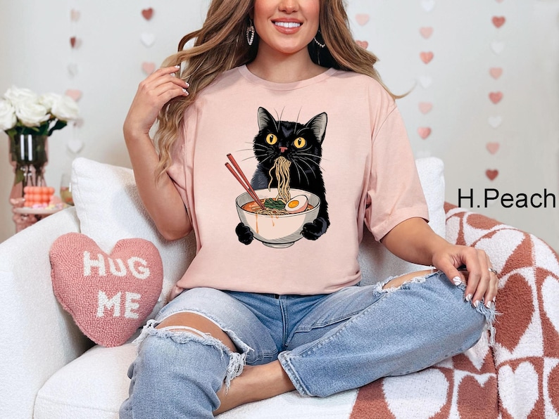 May include: A pink t-shirt with a black cat eating ramen noodles. The cat is holding a bowl of ramen with chopsticks. The text "Hug Me" is on a pink heart-shaped pillow in the background.