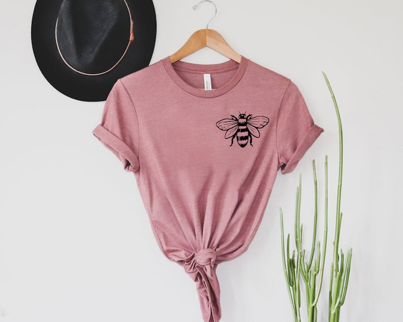 May include: A dusty rose short-sleeve t-shirt featuring a black graphic of a bee on the upper left chest area. The shirt has a crew neckline and is displayed on a wooden hanger, with the bottom hem tied into a knot at the center front.