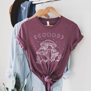 May include: A maroon t-shirt with a white graphic of mushrooms and a crescent moon phase design. The shirt has a knotted front.