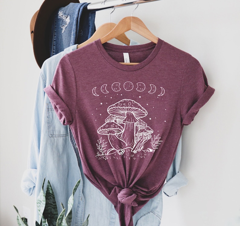 May include: A maroon t-shirt with a white graphic of mushrooms and a crescent moon phase design. The shirt has a knotted front.