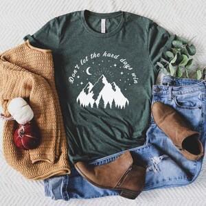 以下が含まれることがあります： 「Don't let the hard days win」の文字と山のグラフィックが入った濃い緑色のTシャツ。ベージュのセーター、青いジーンズ、茶色のアンクルブーツも写っています。