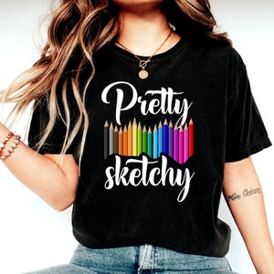 以下が含まれることがあります： 白い文字で「Pretty Sketchy」と書かれた黒いTシャツ。テキストの上に、色とりどりの鉛筆が並んでいます。