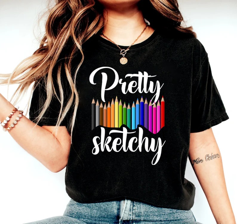 以下が含まれることがあります： 白い文字で「Pretty Sketchy」と書かれた黒いTシャツ。テキストの上に、色とりどりの鉛筆が並んでいます。