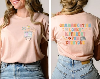 May include: A pink t-shirt with a graphic design featuring a keyboard, a heart, a flower, and a hand sign. The text "Suzy" is printed below the keyboard. The back of the shirt has the text "COMMUNICATION LOOKS DIFFERENT FOR EVERYONE" printed in rainbow colors.