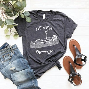 May include: A dark gray t-shirt with a white graphic of a skeleton lying in a coffin with a drink in its hand. The text "NEVER BETTER" is printed below the graphic.