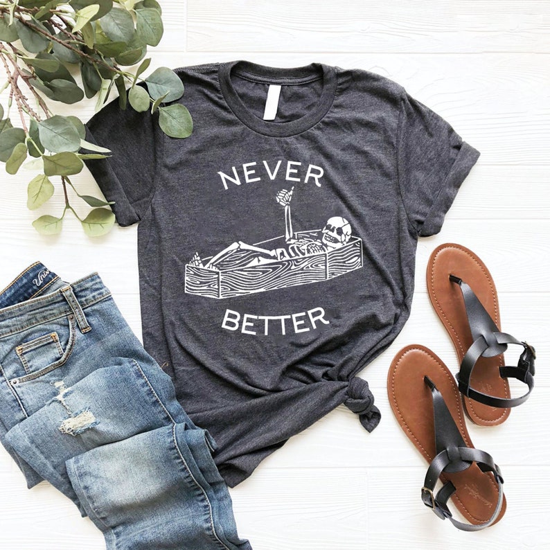 May include: A dark gray t-shirt with a white graphic of a skeleton lying in a coffin with a drink in its hand. The text "NEVER BETTER" is printed below the graphic.