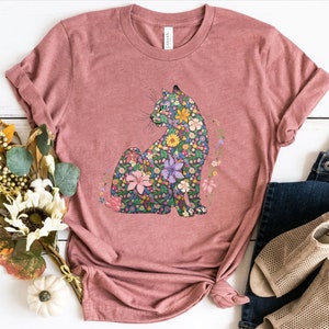 May include: A dusty rose t-shirt featuring a floral cat design. The cat is composed of various colorful flowers and foliage. The shirt is styled with rolled-up sleeves and a knotted hem, paired with blue jeans and beige sandals.