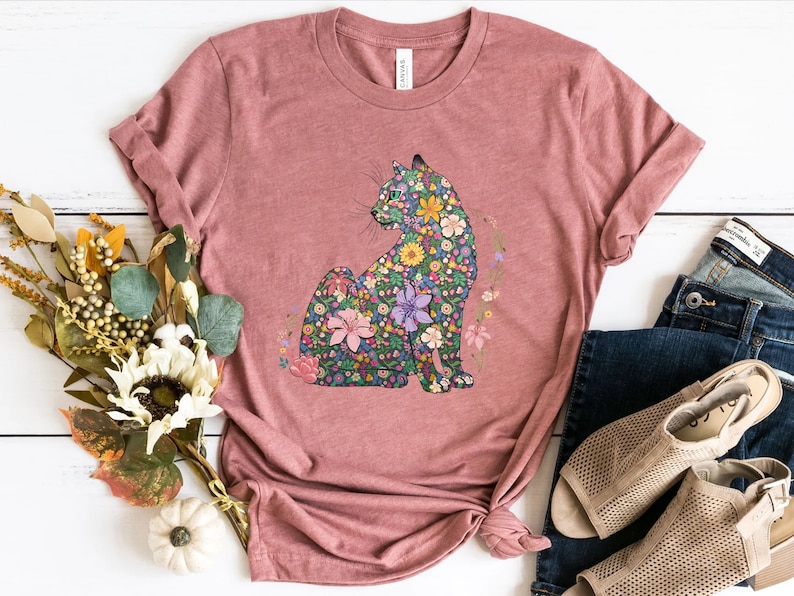May include: A dusty rose t-shirt featuring a floral cat design. The cat is composed of various colorful flowers and foliage. The shirt is styled with rolled-up sleeves and a knotted hem, paired with blue jeans and beige sandals.